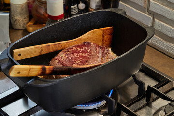 Wall Mural - Piece of meat is fried with onions in wine sauce in rectangular pan over fire on gas stove. Wooden spatulas