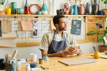 Aufkleber - Young pensive black man in workwear using digital tablet while sitting by table in workshop and scrolling through online information