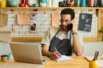 Aufkleber - Young serious clerk of small earthenware shop talking to client by mobile phone while sitting by table in front of laptop in his workshop