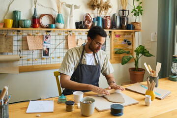 Aufkleber - Young multi-ethnic man in workwear flattening clay for creating new handmade earthenware item while sitting by workplace