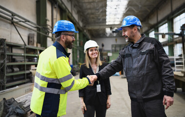 Wall Mural - Engineer and industrial worker in uniform shaking hands in large metal factory hall and talking.