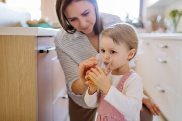 Young mother giving glass of water to drink to her little daughter in kitchen at home.