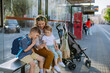 © Halfpoint - Young mother with little kids waiting on bus stop in city, scrolling on mobile phone.