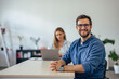 © bnenin - Portrait of a smiling man with glasses on, posing for the camera while a female working in the background.