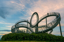 Walkable Tiger & Turtle Roller Coaster Sculpture On Magic Mountain With Some People In Motion Blur Against A Cloudy Sunset Sky, Art Installation And Landmark In Angerpark Duisburg, Germany, Copy Space