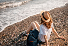 Young Woman In A White T-shirt And Hat Sitting On A Pebble Beach And Looking At The Sea