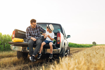 father and son sits on truck trunk outdoor on field . they enjoying time together