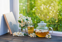 Hot Herbal Tea In Glass Teapot, Cup And Beautiful Bouquet Of Jasmine Flowers On Windowsill At Summer Day Near Garden