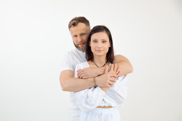 Bearded man behind and woman, happy family in light outfit stand together, look at camera, hugging on white background.