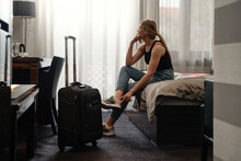 Woman Taking Off Footwear In Sunny Hotel Room On The Bed. Tourist Relaxing On Hotel Room After Travelling With Suitcase. Happy Female Having Rest After Long Trip With Language Dark Silhouette 