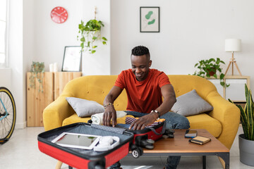 Young man packing travel bag for summer vacation - African male preparing suitcase before going on holidays