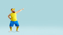 Funny Plump Man In Sportswear Dancing Isolated On Blue Copy Space Background. Happy Cheerful Fat Guy In Yellow Top And Blue Shorts Pointing To Copyspace On Right Side. Sport, Fitness Workout Concept