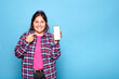 © Lux Images - Young curvy latina woman in a plaid shirt holding and pointing at her mobile phone, isolated against a blue background.