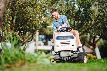 Wall Mural - Gardener driving a riding lawn mower in a garden. Professional landscaper using tractor at mowing lawn.