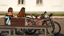 Two Girls Are Sitting On A Bench Outside After A Bike Ride. There Are Bicycles Nearby. Selective Focus.