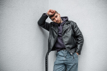 Wall Mural - Fashionable hipster handsome man model with hairstyle in fashion rock leather jacket with jeans and hoodie stands and poses near a white wall on the street