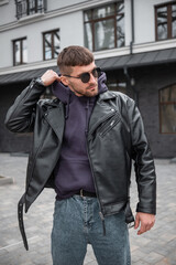 Wall Mural - Fashionable handsome hipster man with sunglasses in a trendy rock black leather jacket and hoodie walking in the city and wearing a hoodie. Men's streetwear style