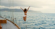 Young Woman In A White Bikini Excitedly Jumping Off A Yacht Into The Ocean. Woman Sailing Around Italy Jumping Off A Yacht To Swim In The Ocean. Woman In White Bikini Jumping Into The Sea To Swim