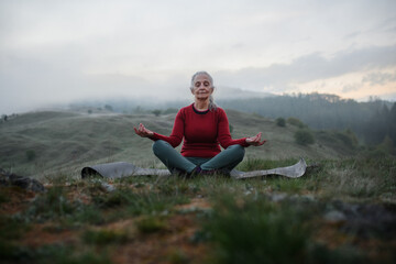 Senior woman doing breathing exercise in nature on early morning with fog and mountains in background.