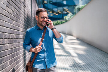 Smiling businessman talking on smart phone while walking by wall on footpath