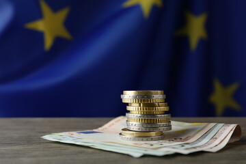 Stack of coins and banknotes on wooden table against European Union flag, space for text