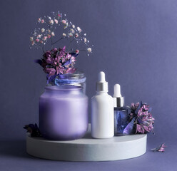 Purple beauty product setting with cosmetic serum bottles, candle and flowers. Healthy skin care products for facial treatment. Front view with copy space.