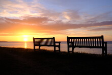 Silhouette Of Two Benches Set Against A Beautiful Seaside Sunset.