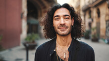 Young Attractive Italian Guy With Long Curly Hair And Stubble Looks Into The Camera And Smiles At Old Buildings Background. Stylish Man With An Earring In His Ear And Lot Of Chains