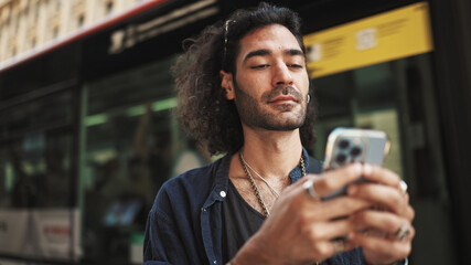 Young attractive italian guy with long curly hair and stubble is using mobile phone. Stylish man with writes message on smartphone