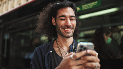 Young attractive italian guy with long curly hair and stubble is using mobile phone. Stylish man with writes message on smartphone