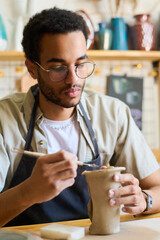 Aufkleber - Serious guy in eyeglasses and apron using handtool while creating clay jug or vase for collection of earthenware items for sale