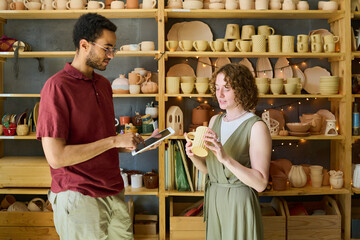 Aufkleber - Young female holding handmade clay mug during discussion of its ornament or other characteristics with colleague against display