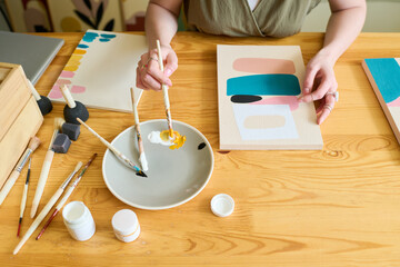 Aufkleber - ABove angle of hands of young female artist mixing colors on plate while sitting by wooden table and painting abstract picture on paper