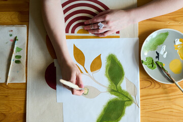 Aufkleber - Above angle of hands of young female artist with paintbrush painting green leaves on paper while sitting by table in workshop
