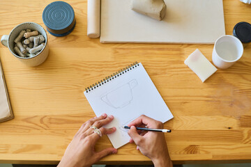 Aufkleber - Above angle of hands of young craftsman or artisan drawing sketch of new earthenware item in notepad while working by wooden table
