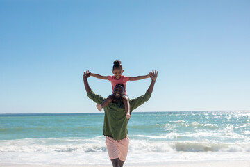 Cheerful african american father carrying daughter on shoulders at beach on sunny day, copy space