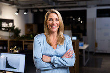 Portrait Of Cheerful Caucasian Businesswoman With Arms Crossed Standing In Modern Office At Night