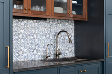 A Kitchen Sink With A Beautiful Pattern Tiled Backsplash With A Chrome Faucet, Black Granite Countertops, And Surrounded By Blue And Wood Cabinets.