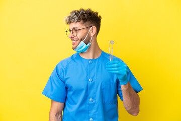 Wall Mural - Dentist caucasian man holding tools isolated on yellow background looking side