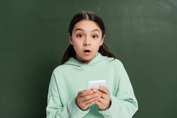 Wall Mural - Shocked schoolkid holding smartphone near chalkboard at background.