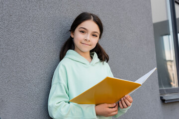 Wall Mural - Smiling schoolkid holding notebook and looking at camera near building outdoors.