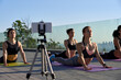 © insta_photos - Fit sporty young women doing yoga surya namaskar pose live stream online social media training record video blog remote group fitness tutorial vlog workout class outdoor on phone camera on tripod.