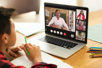 Canvas Print - Focused asian boy with book and pencil studying online through video call over laptop at home