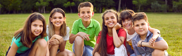 Wall Mural - Happy children sitting on green grass and smiling. Cheerful little kids enjoying their school break, spending time in a summer camp, making new friends, and playing outside. Group portrait. Banner