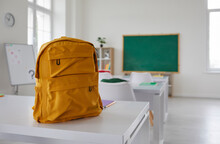 School Classroom. New School Bag On A Student's Desk In The Classroom. Big Yellow Canvas Backpack Placed On The Table In A Large Modern Schoolroom With A Chalkboard. Back To School Concept