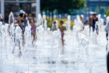 Selective Focus On The Water Jets Of The City Fountain. Against A Blurred Background, People Bathe In The Jets Of A Fountain Splashing From The Ground During Hot Weather In The Summer In The City.