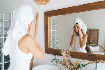 Young beautiful female with towel on head smiling to mirror reflection  standing in the bathroom at home. 30 years old happy woman doing daily morning rituals and cleansing. Enjoying healthy skin care