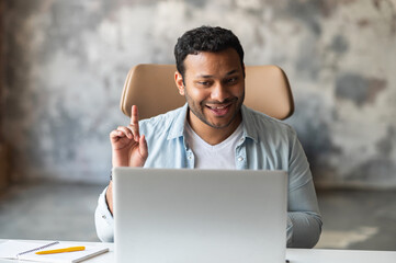 Wall Mural - Front view of indian male office employee talking via video connection while sitting at the desk. A guy using laptop for video call in office space, talking on the distance with colleagues or clients