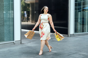 Happy confident smiling plus size curvy young woman with shopping bags walking on city street near shop windows