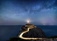 Night Time Image With Milky Way Stars And Illuminated Road With Light Trails At The Far De Formentor Lighthouse On Mallorca, Spain